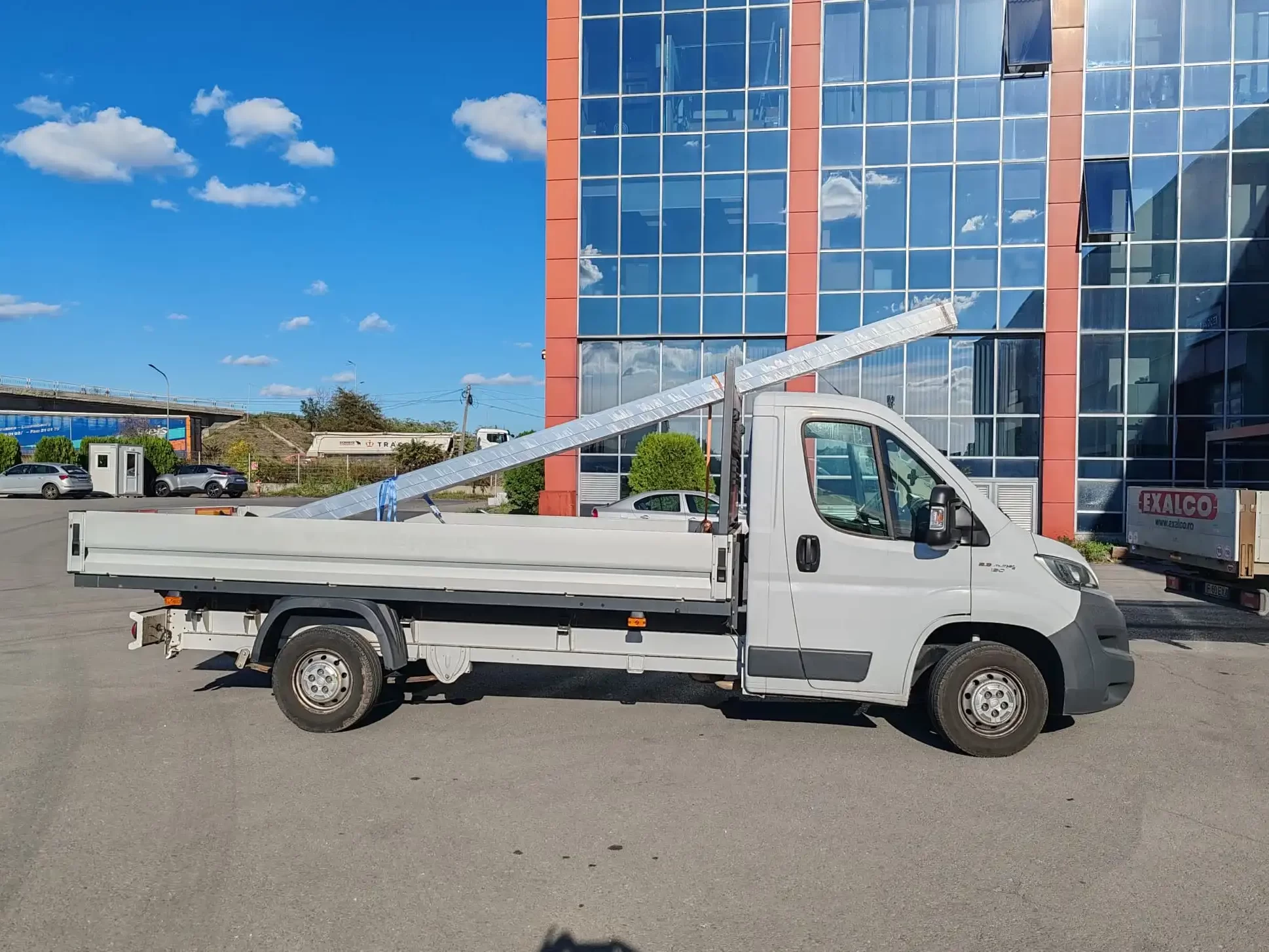 Autoutilitara Fiat Ducato de culoare alba , pe care sa scrie textul: 'Ovi Trans ' , cu usile din spate deschise , iar in spatele ei se vede cum un stivuitor incarca un palet cu marfa